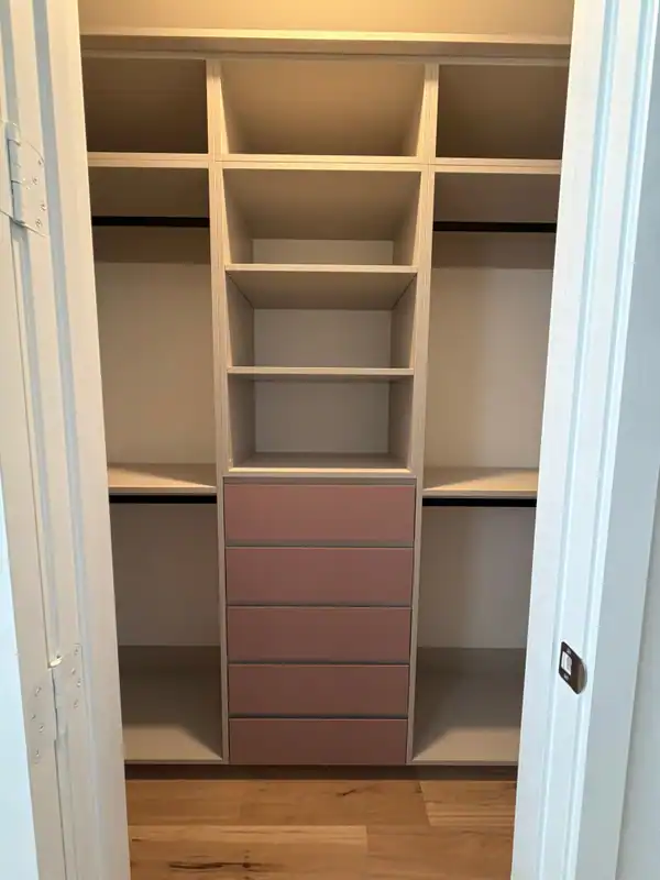Reach-in closet with cream shelving, black hanging rods on both sides and a centre stack of five mauve-pink drawers above a wood-grain floor
