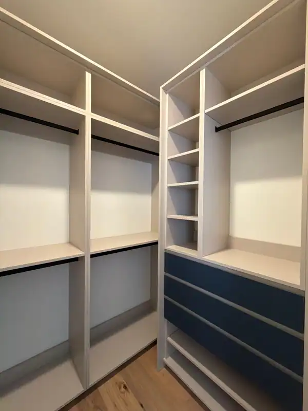 Empty corner walk-in closet with white open shelving, black hanging rods and a bank of navy blue drawers, over a light wood-grain floor