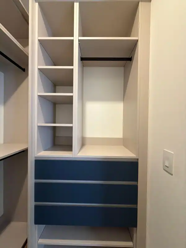 White closet unit with open shelf compartments, a black hanging rod and three navy blue drawers below, beside a wall light switch