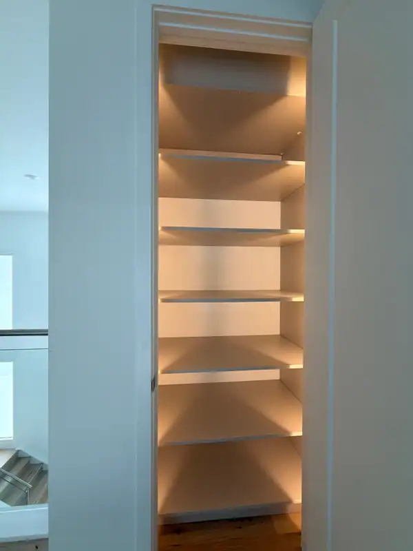 Tall built-in niche with six white shelves and warm LED strip lighting, set into a white wall beside a stair landing