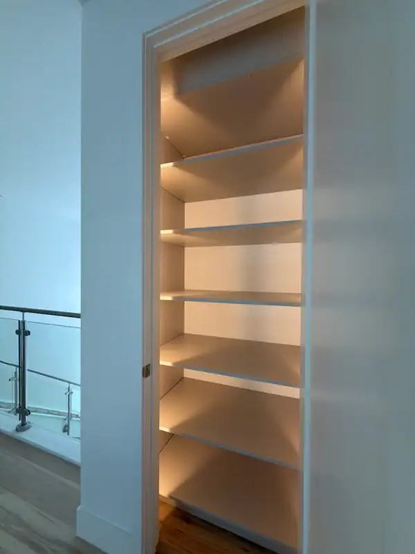 Tall recessed shelving niche with six white shelves lit by warm LED strips, next to a glass-panel railing and a wood-grain floor