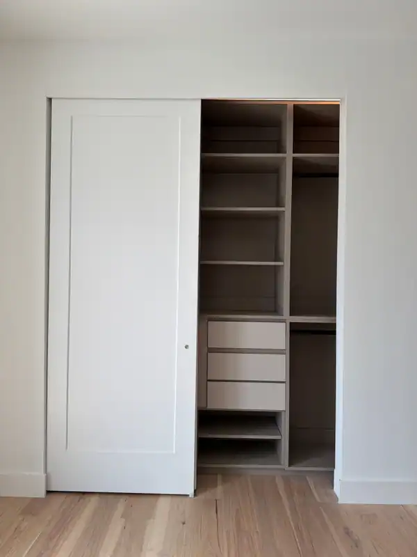 Closet with a white sliding panel door pushed open to show greige shelving, three drawers and hanging rods, over a light wood-grain floor