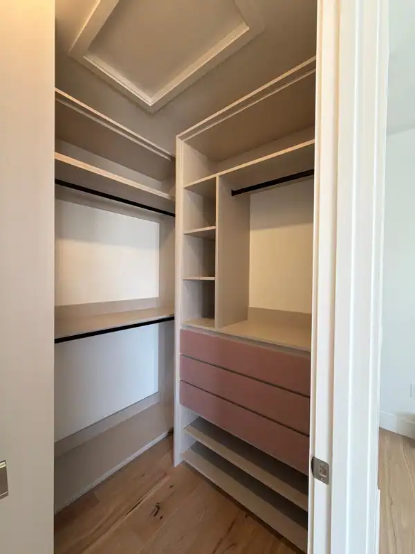 Walk-in closet corner with pink drawer fronts, greige open shelves, black hanging rods and a recessed tray ceiling above a wood-grain floor