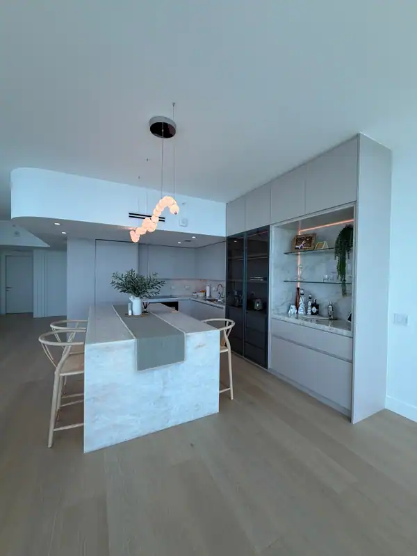 Kitchen with a veined-stone waterfall island, woven-seat stools, a wavy bubble pendant, and dark glass-front tall cabinets beside a lit display niche