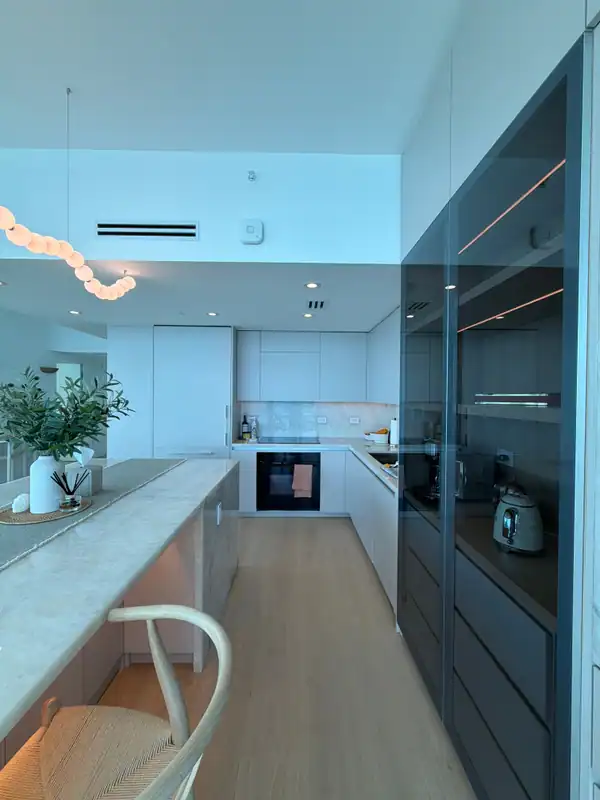 Kitchen with white flat-panel cabinets, a built-in oven, a stone-top island with a woven-seat stool, and a tall smoked-glass pantry cabinet
