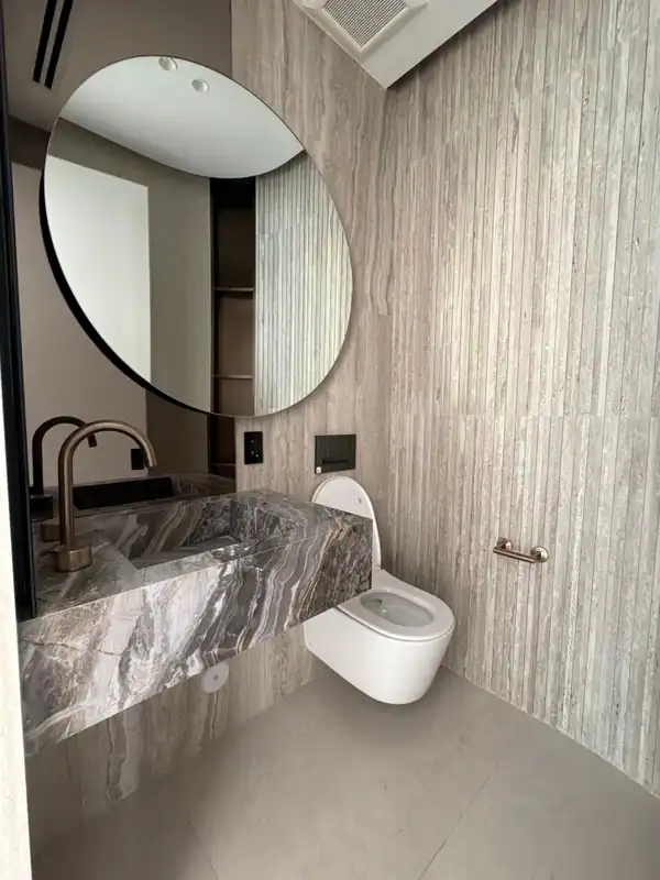 Bathroom with a large round black-framed mirror, vertical striated stone-look wall tile, a floating veined stone vanity, and a wall-hung toilet