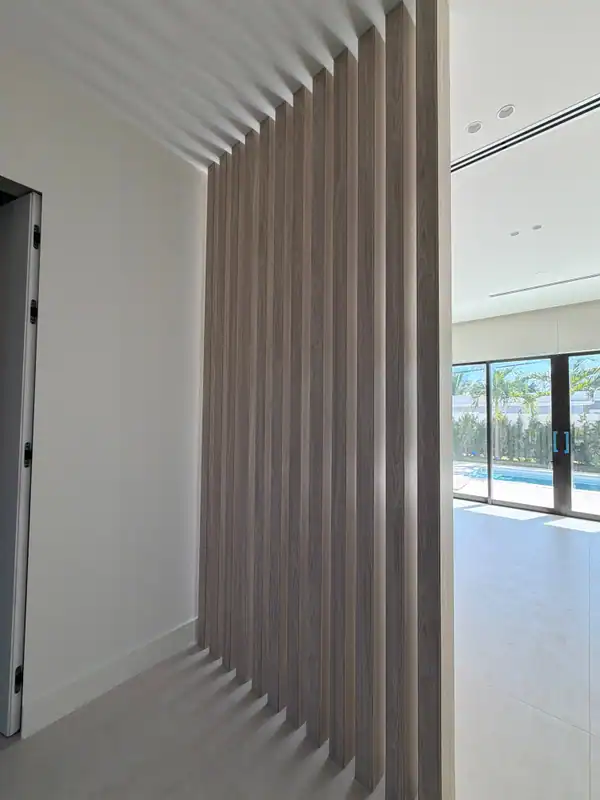 Taupe wood-grain vertical slat screen with staggered ends against a white wall, glass sliders to a pool and palms at right, light tile floor