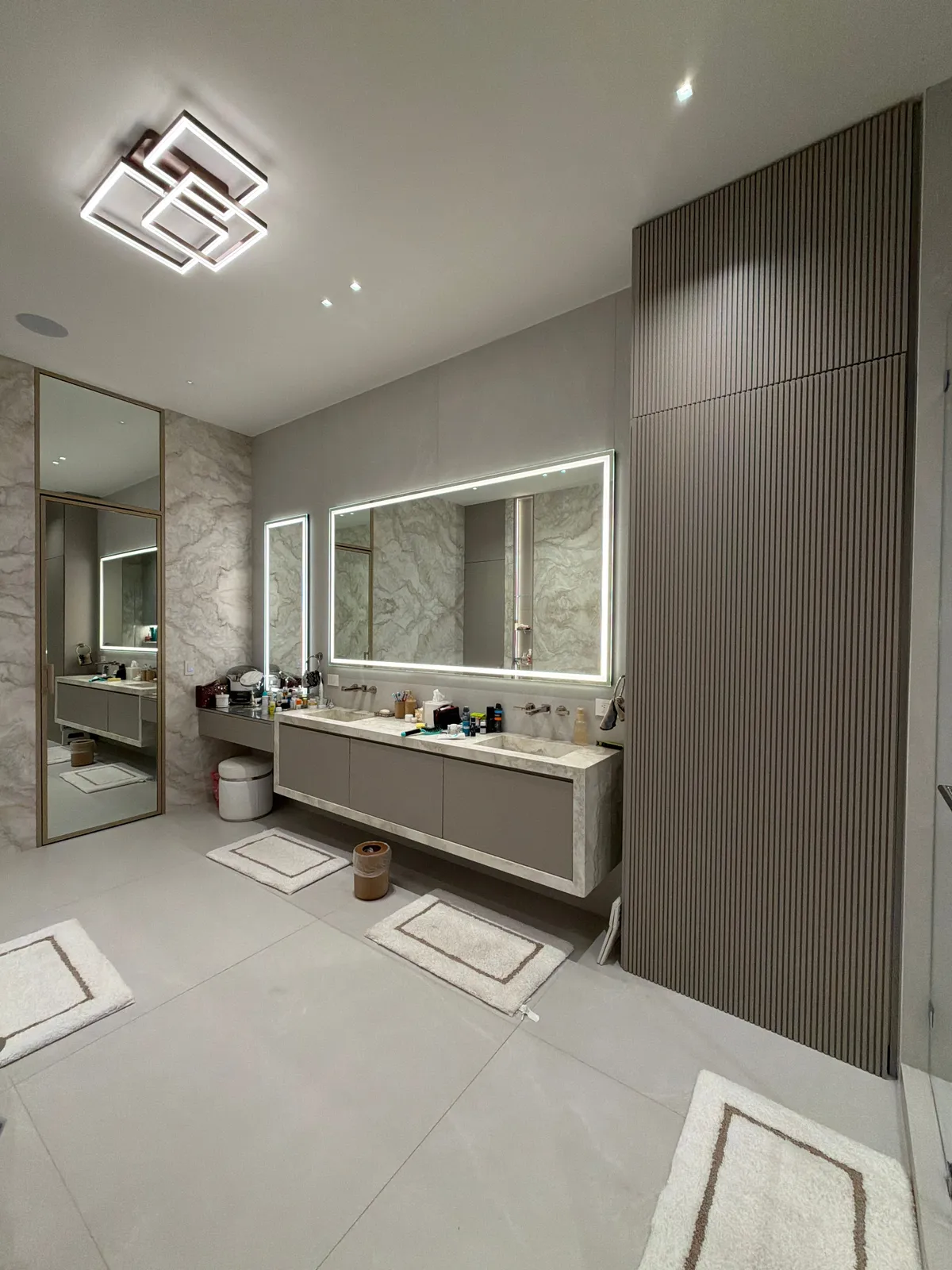 Bathroom with a floor-to-ceiling fluted wood-look wall panel, a taupe floating vanity with stone-look top, backlit LED mirrors, and bath mats