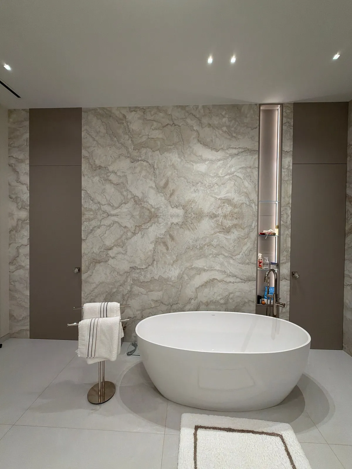 Freestanding oval tub before a veined stone slab wall flanked by taupe flat-panel cabinetry, with a lit niche shelf and a standing towel bar