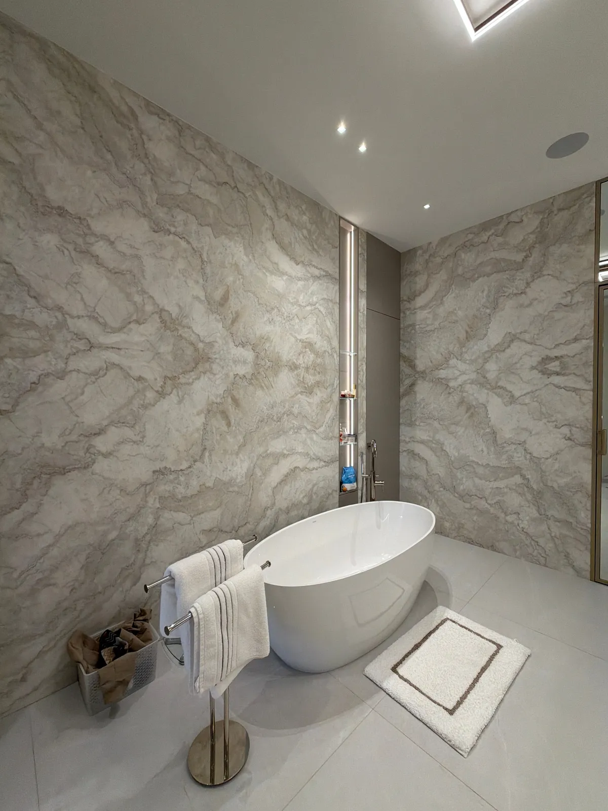 Freestanding oval tub against veined stone slab walls, with a lit recessed niche, a standing towel bar with white towels, and a bath mat