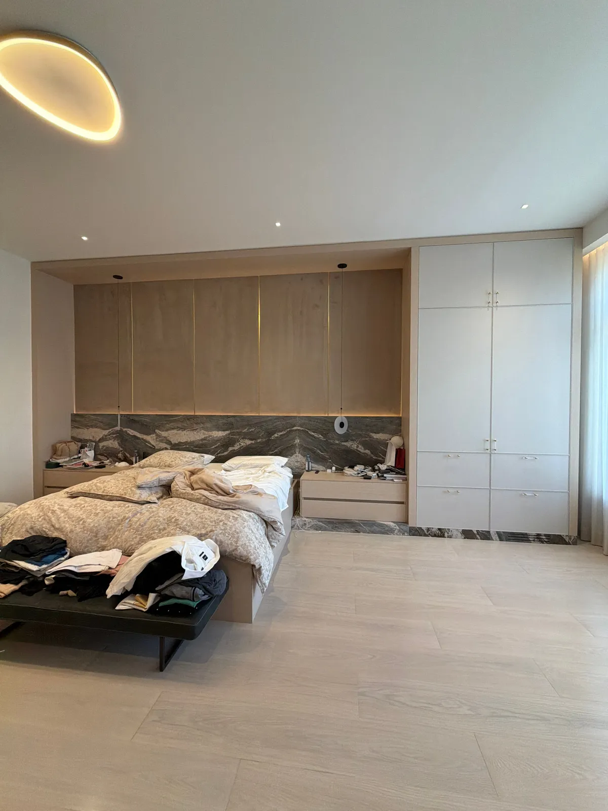 Bedroom with a wood-grain overhead cabinet run over the bed, against a dark veined stone wall, tall white closet unit at right, clothes on a black bench