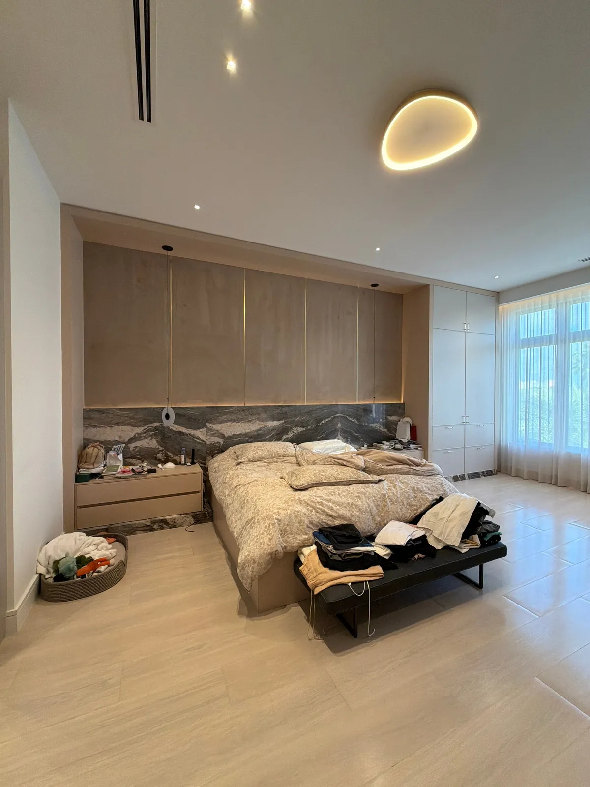 Bedroom with wood-grain overhead cabinets above the bed, set against a dark veined stone wall, tall white closet doors, black bench, light tile floor