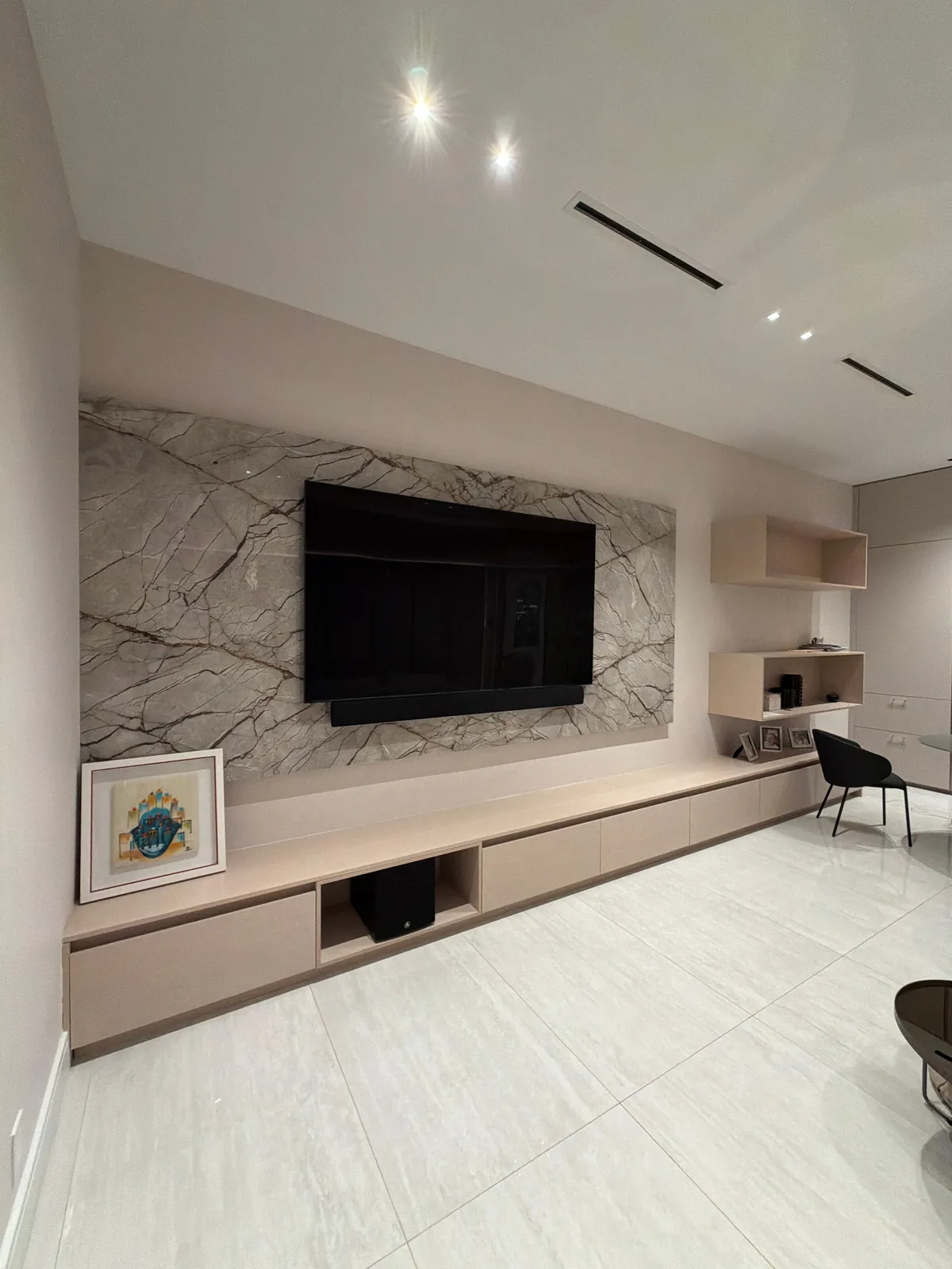 Angled view of a veined stone wall with a mounted TV above a long low wood-grain credenza, framed art leaning at the end and open shelf boxes right