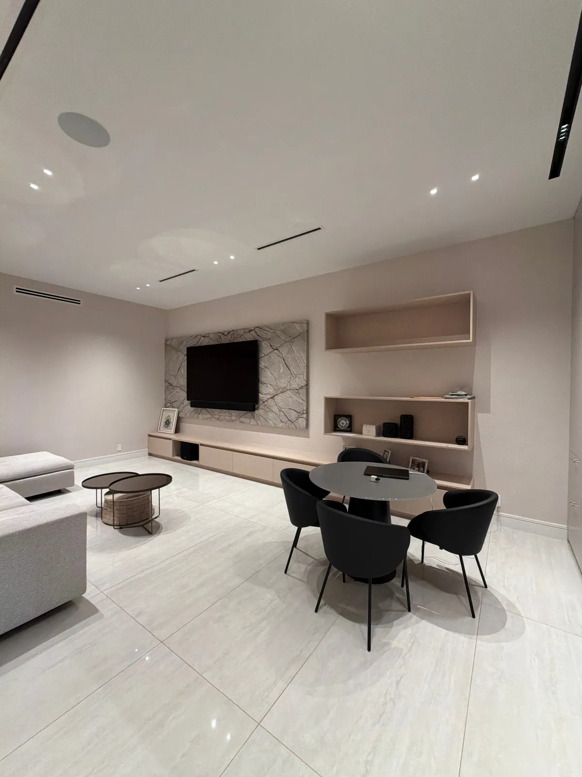 Room with a veined stone wall behind a mounted TV, a long low wood-grain credenza, open wall shelf boxes and a round dark table with black chairs