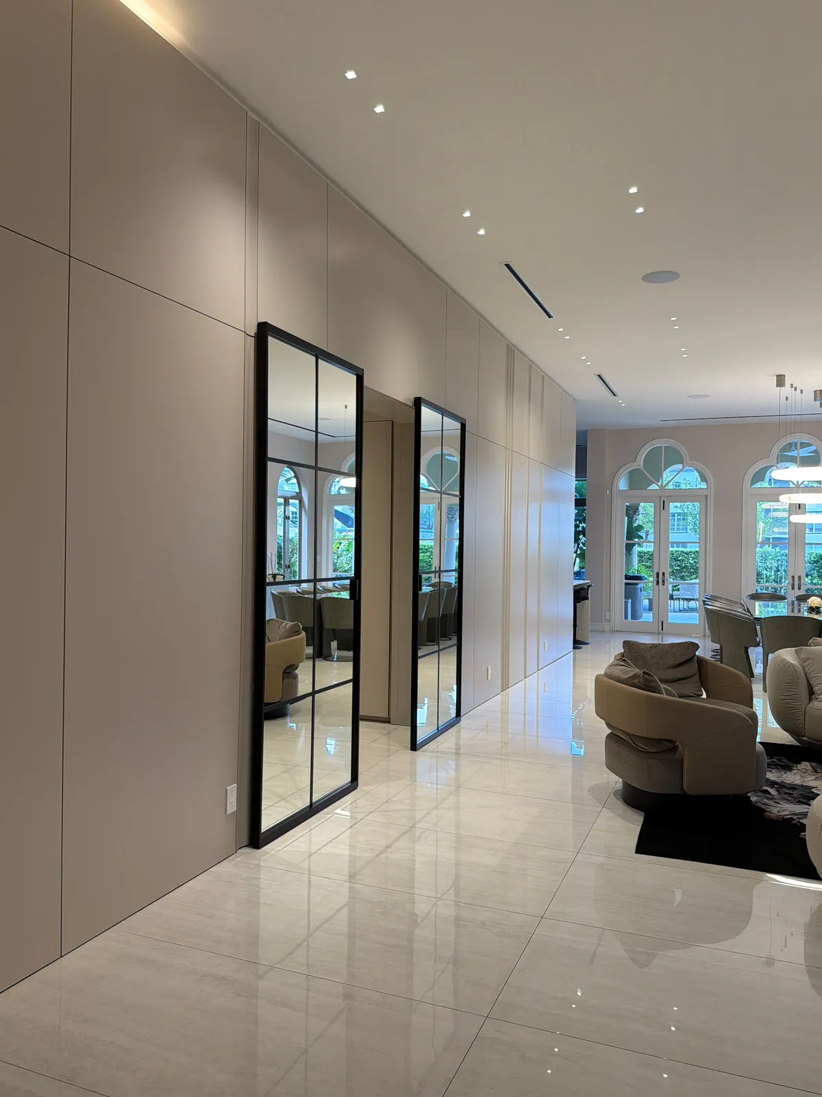 Cream flat-panel wall with two tall black-framed grid-glass doors reflecting arched windows, curved upholstered chairs on polished stone tile
