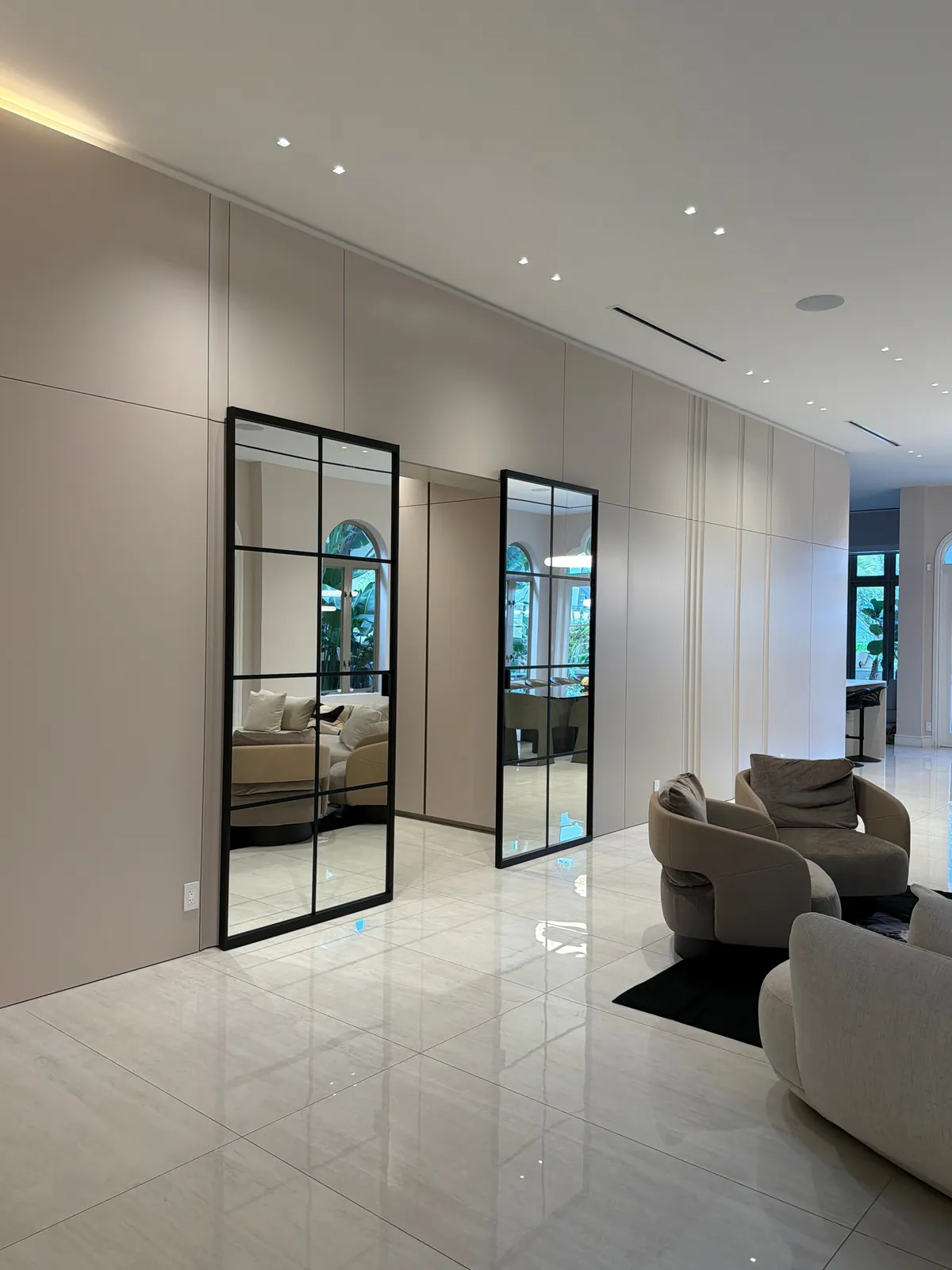 Cream flat-panel wall with two tall black-framed grid-glass doors opening to a living area with upholstered sofas, over a polished stone tile floor