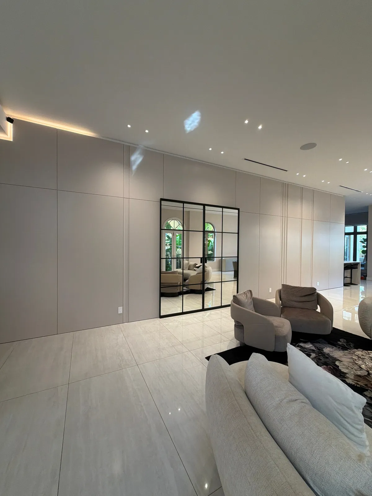 Long flat taupe panel wall with a large black grid-framed mirror, cream sofa and taupe swivel chairs on a dark floral rug, light tile floor