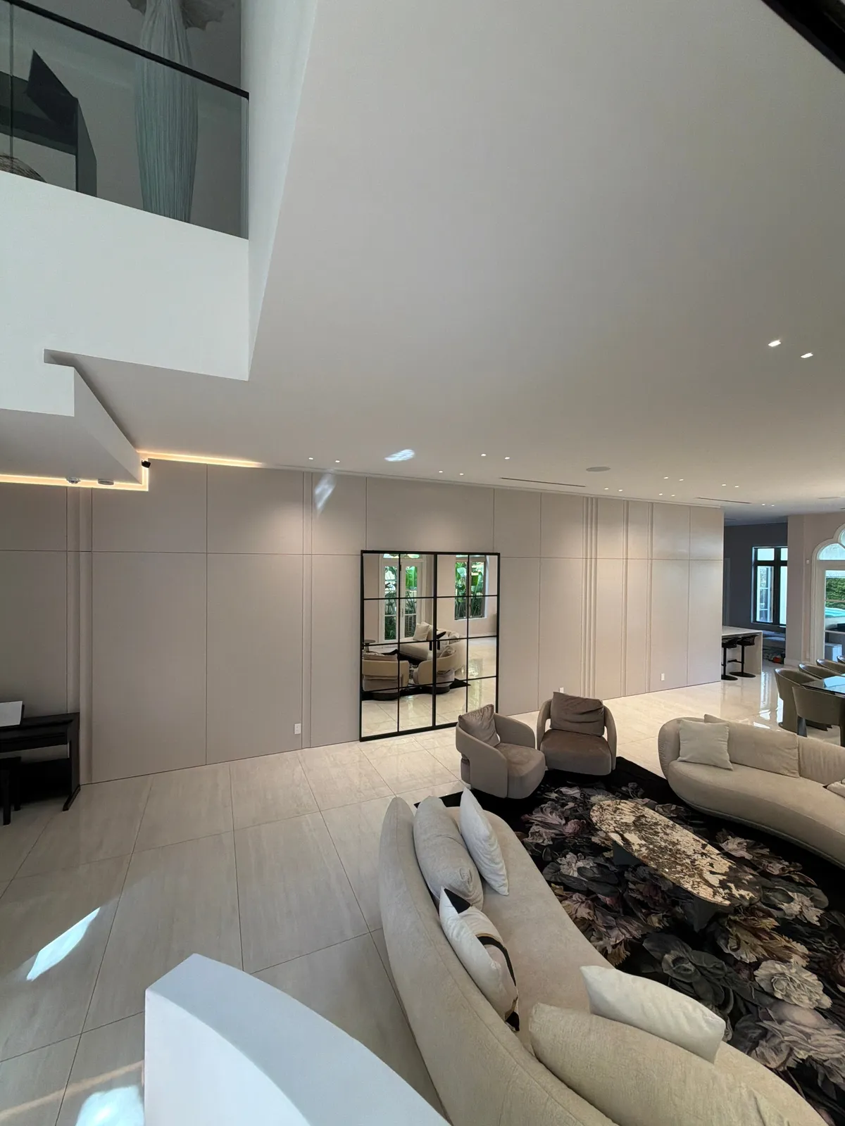 Open room with a long taupe flat-panel wall and black grid-framed mirror, cream curved sofas and floral rug, upper-level glass balustrade above