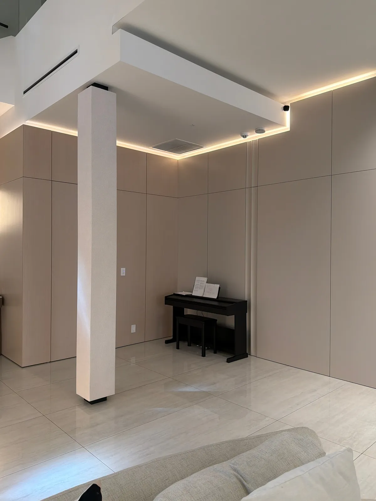 Taupe panel walls meeting at a corner with a white column, cove lighting above, black upright piano and bench, glossy light tile floor