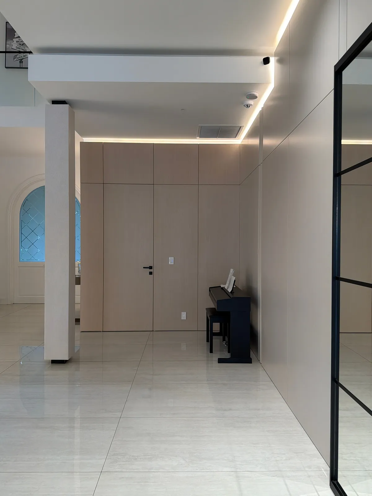 Taupe panel wall with cove lighting and a flush door, black upright piano at the corner, glossy light tile floor, black-framed glass partition at right