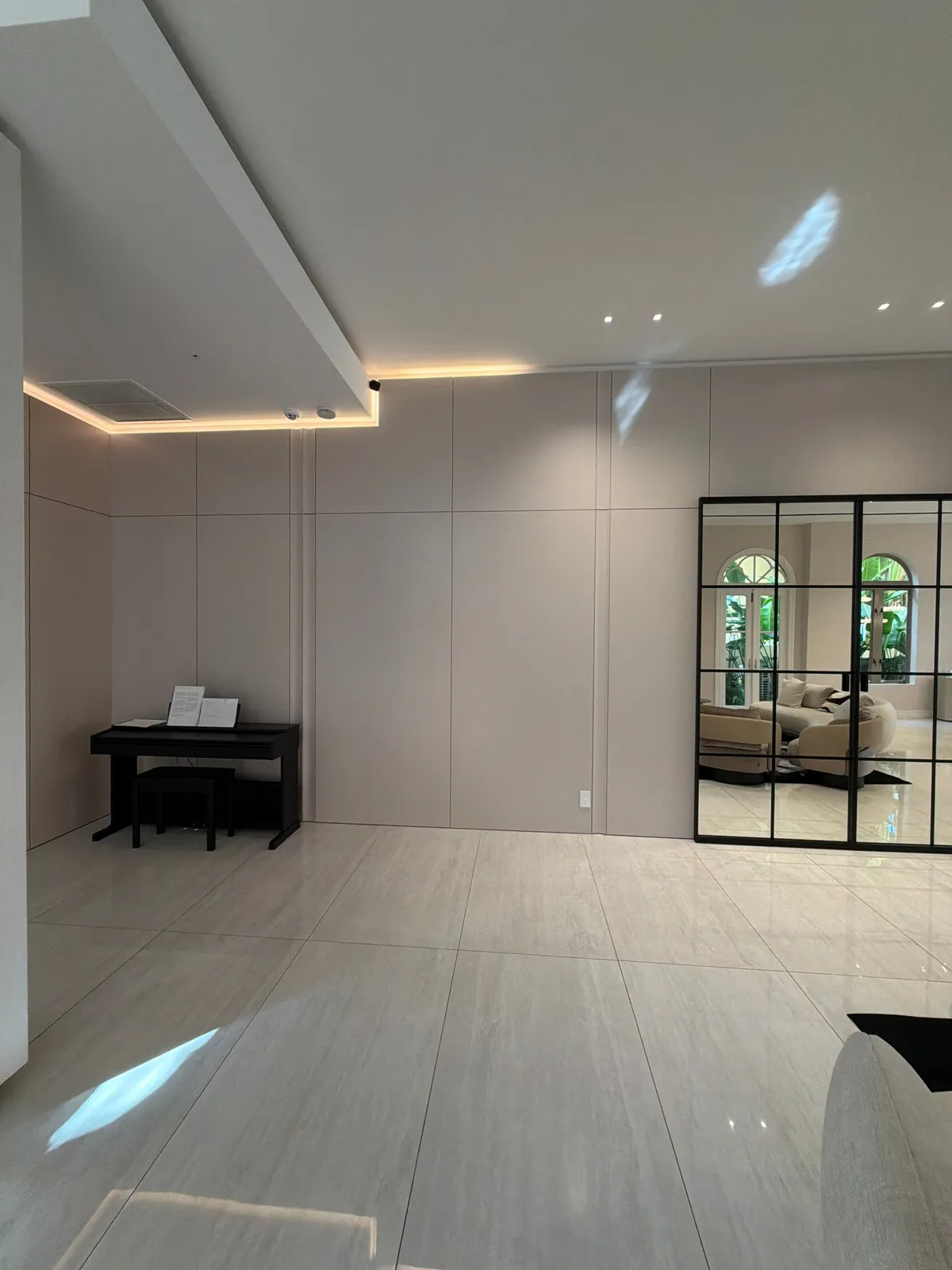 Taupe flat panel wall with cove lighting, a black upright piano in a recess, and a tall black grid-framed mirror reflecting arched windows