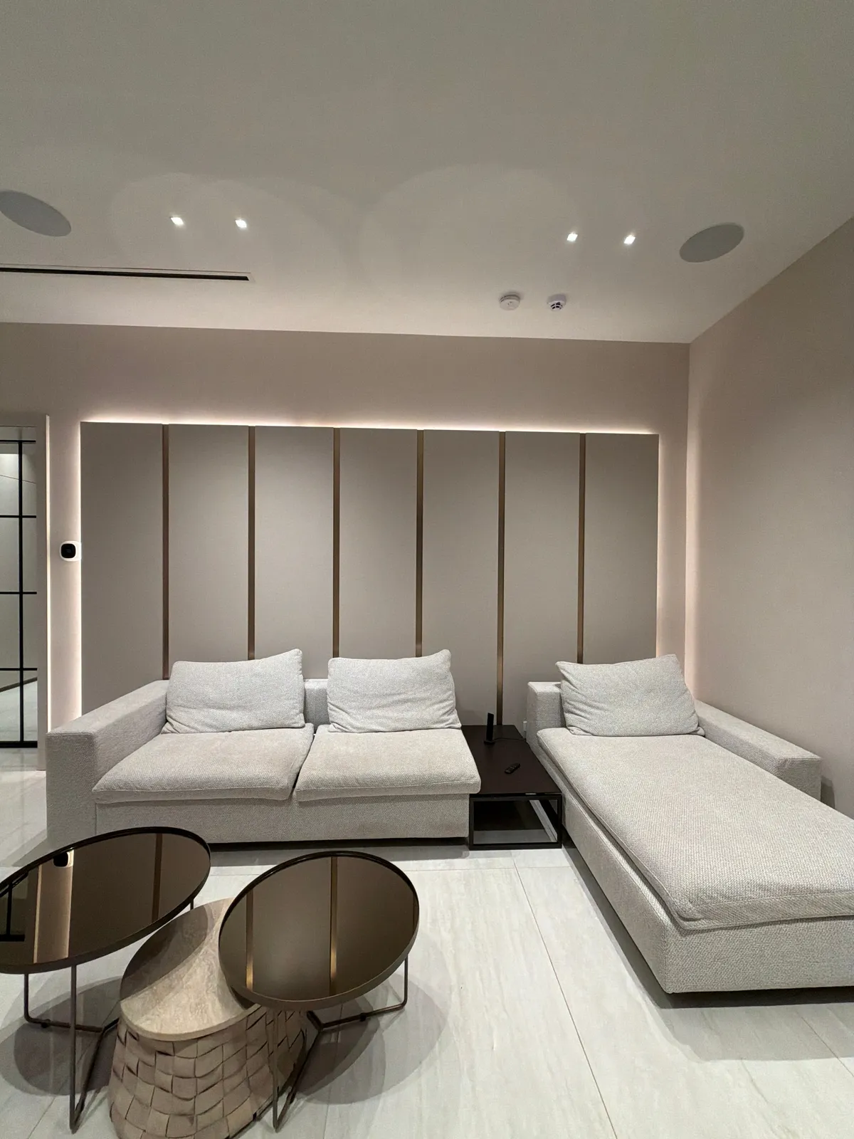 Taupe panel wall with slim vertical metallic reveals and backlit edge glow, light upholstered sofa and chaise, two round dark glass tables on tile