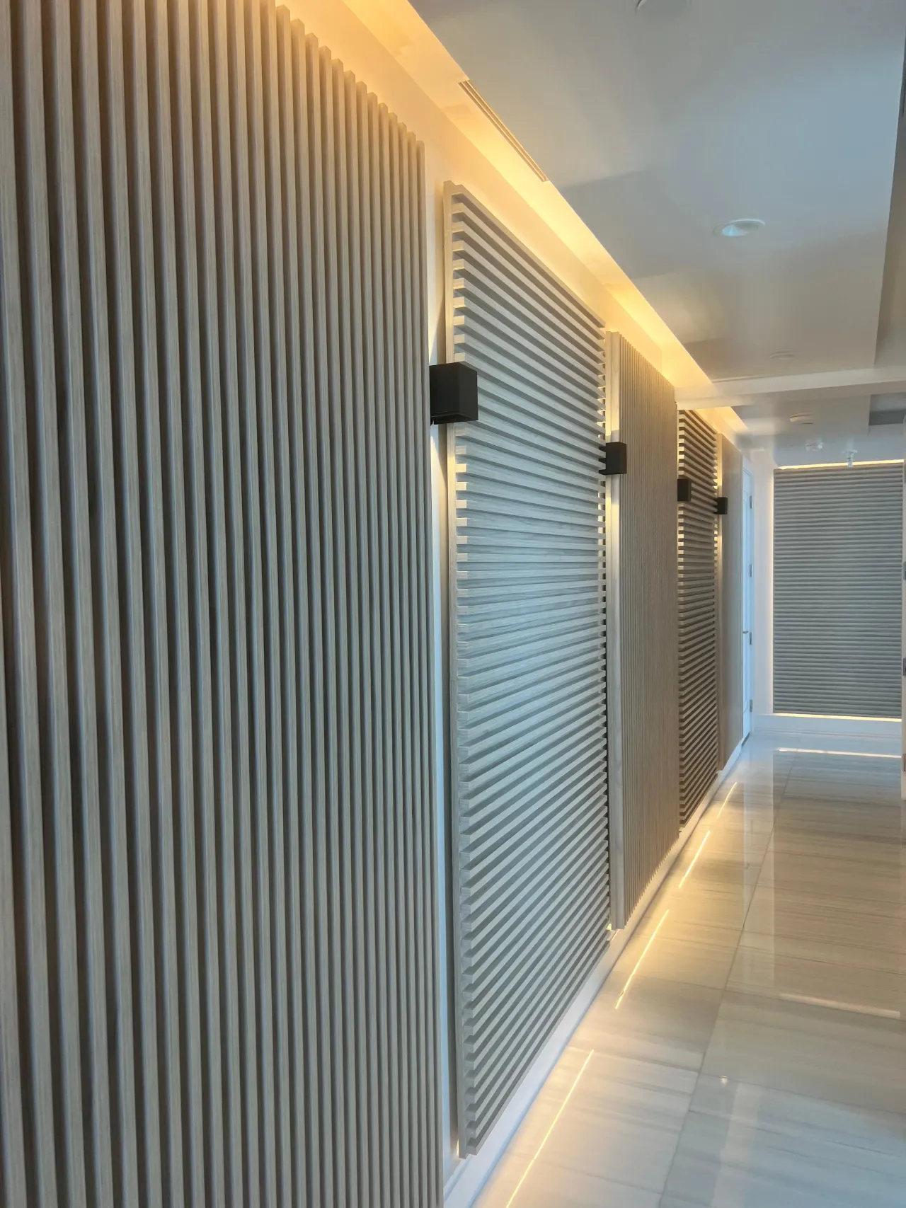 Corridor lined with light wood-grain slat panels alternating vertical and horizontal runs, black box sconces between them, glossy light tile floor