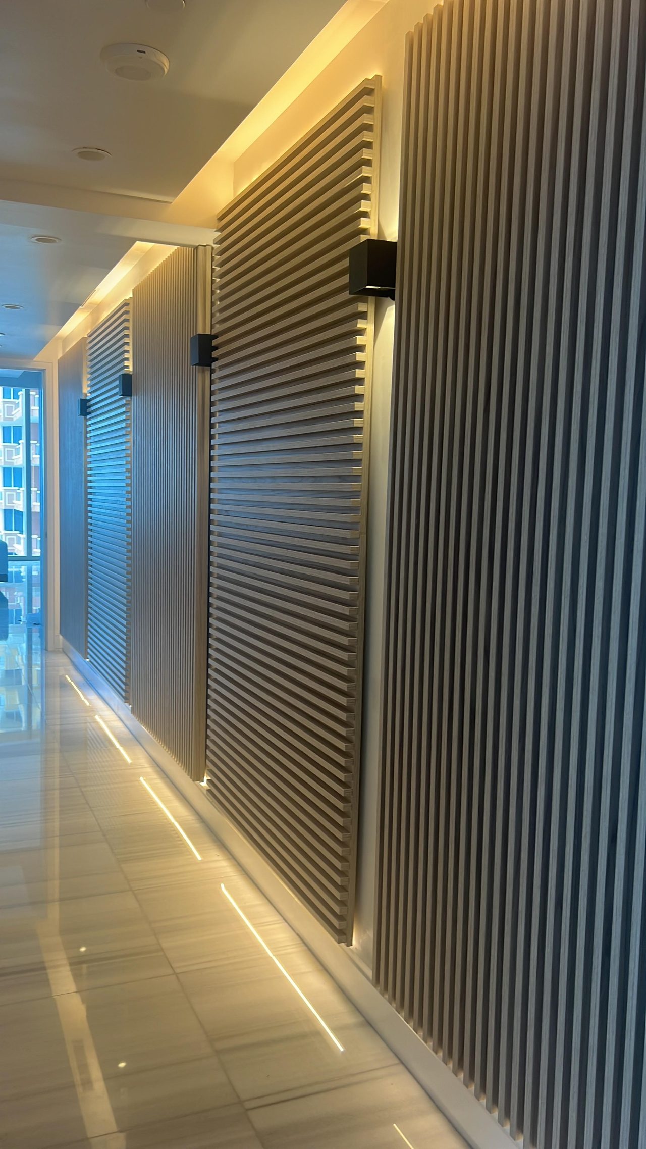 Corridor of light wood-grain slat panels alternating vertical and horizontal sections, surface-mounted black sconces, cove lighting and glossy tile floor