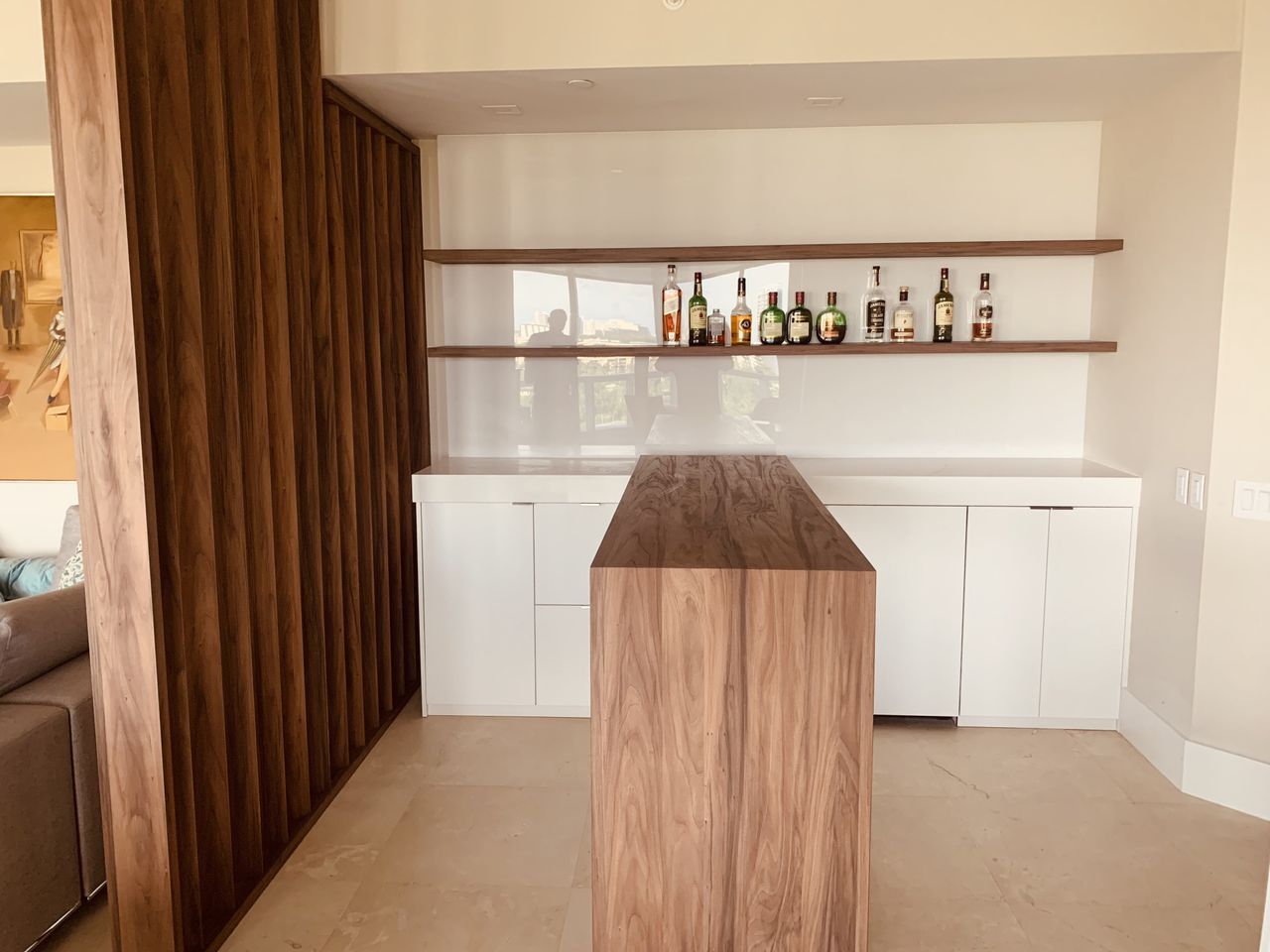 Dark wood home bar with two floating shelves holding liquor bottles over a white cabinet base and a waterfall-edge bar counter