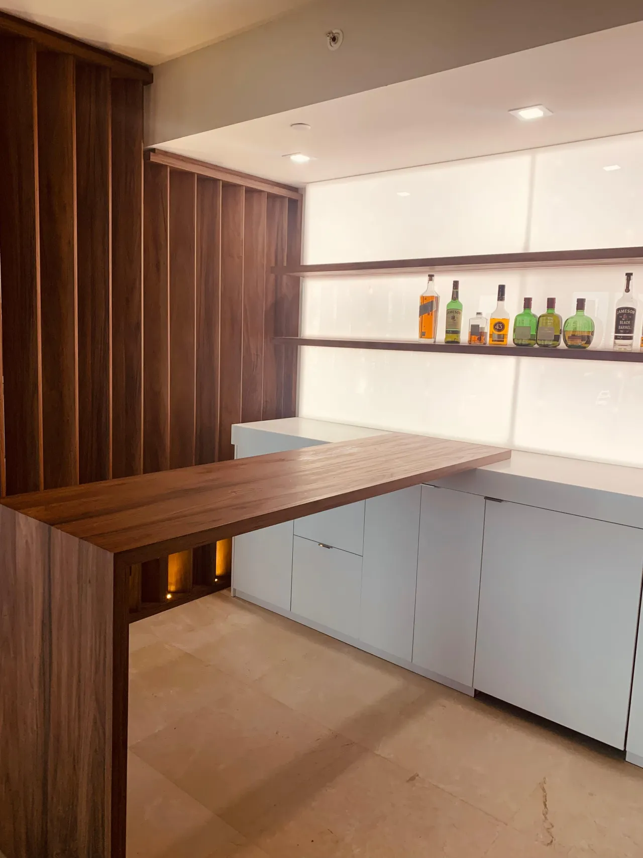 Dark wood home bar with a vertical fluted side wall and a floating open shelf holding liquor bottles against a backlit white panel