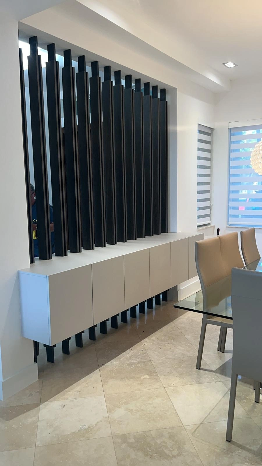 Dark wood slat wall divider set into a ceiling niche above a white floating credenza in a dining area, window blinds visible at right