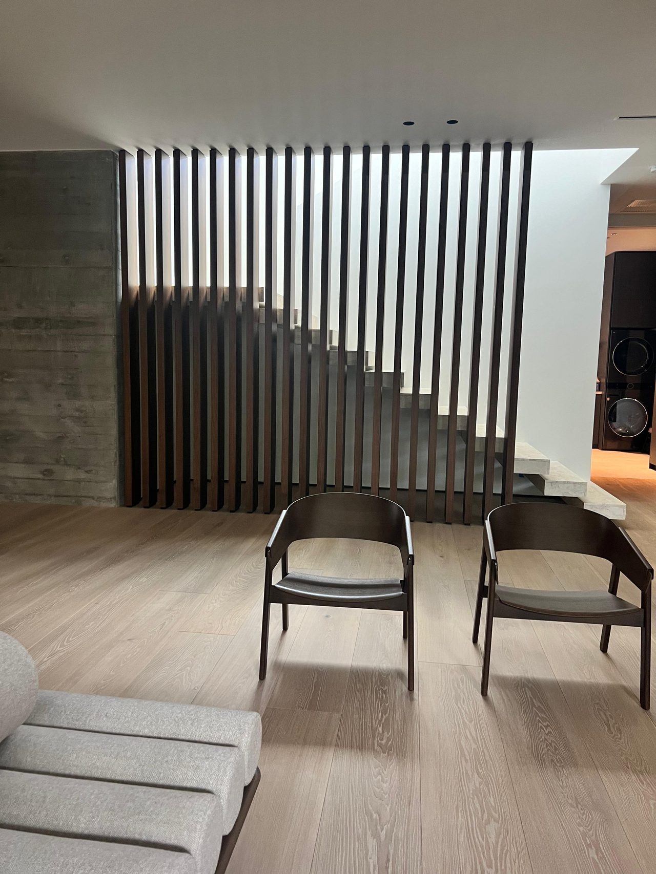 Dark wood vertical slat screen with staggered ends in front of a staircase, board-formed concrete wall at left, two dark wood armchairs on light wood floor
