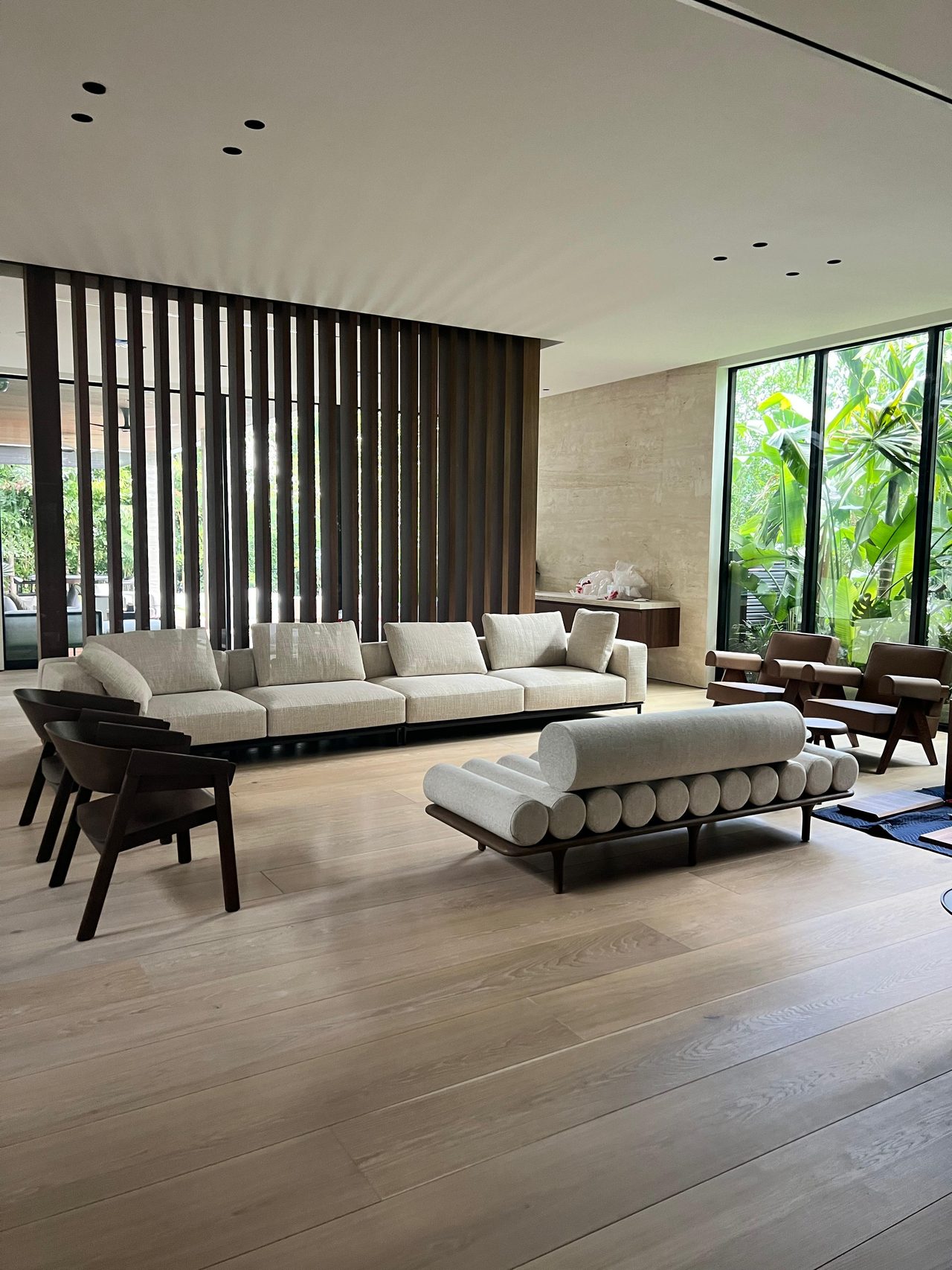Dark wood vertical slat screen along a glazed wall in a living room with a cream sectional, bolster daybed and dark wood armchairs on light wood floor