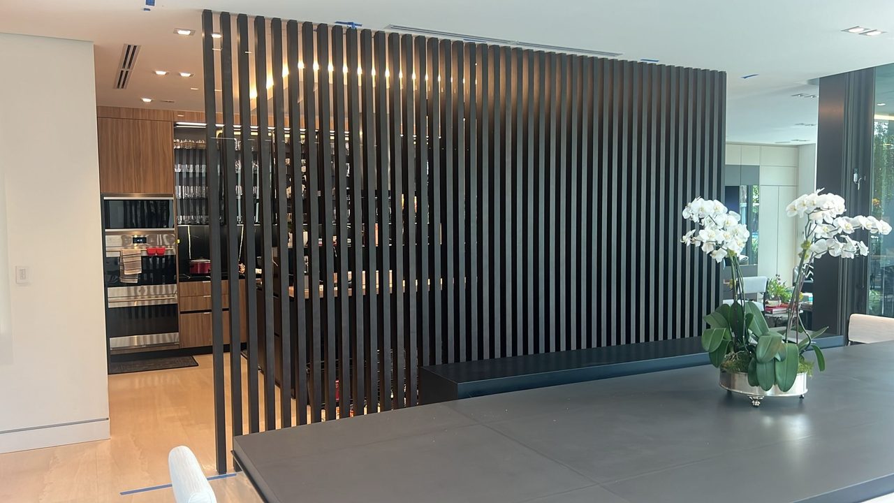 Floor-to-ceiling dark vertical slat screen dividing a kitchen from a dining area, wood-grain cabinetry and stainless ovens beyond, dark stone counter