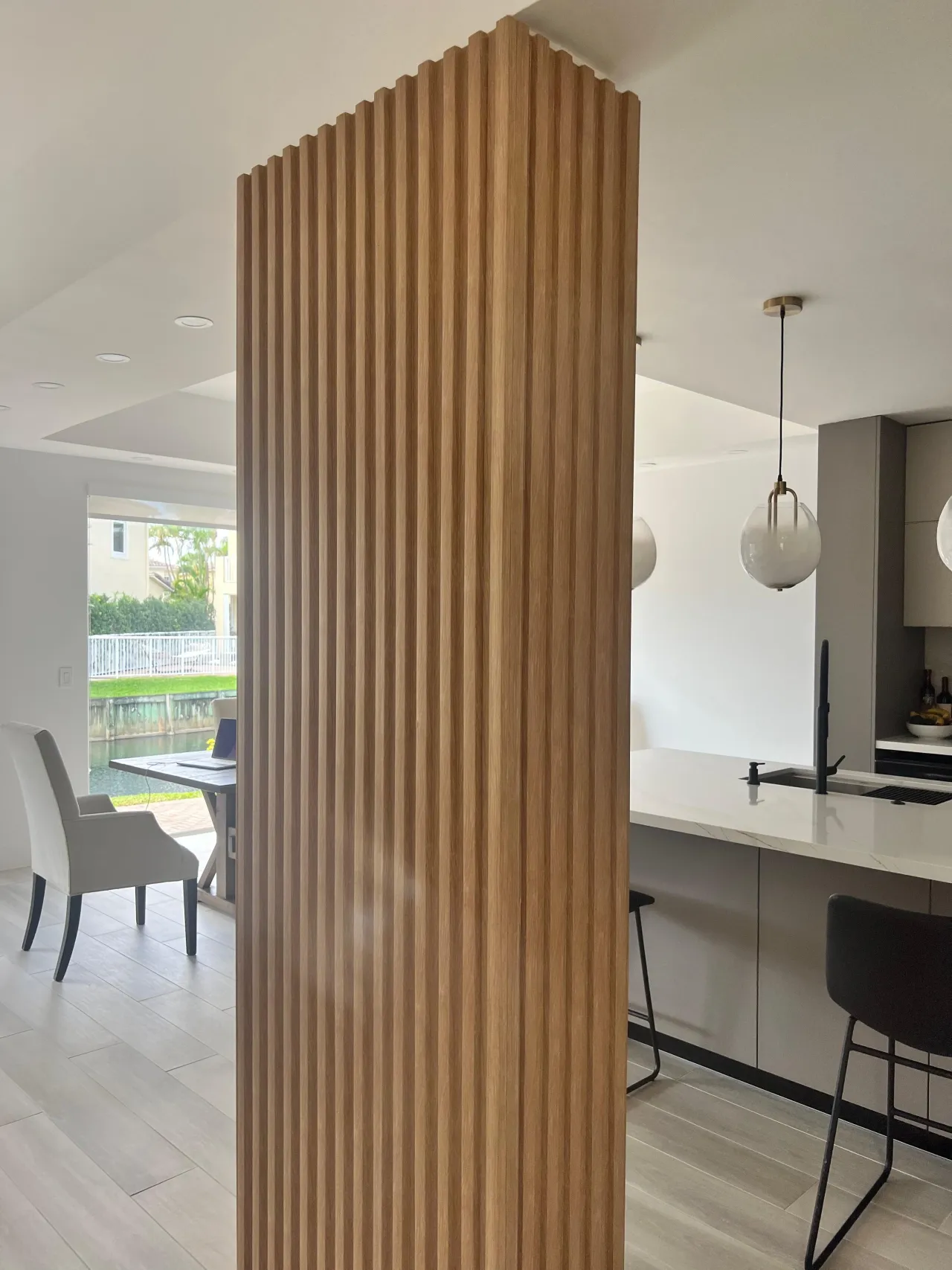 Fluted wood-grain column wrap between a seating area and an open kitchen with white globe pendants, a light stone counter and dark barstools