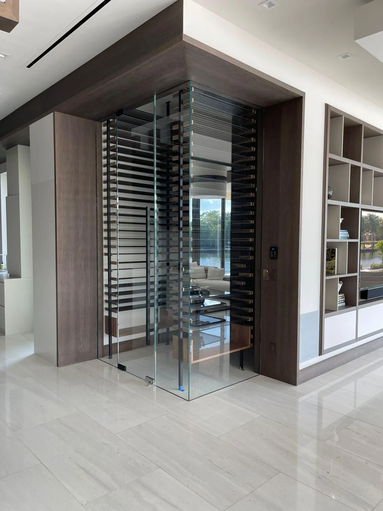 Glass-enclosed wine cellar with dark wood surround and black horizontal wine bottle racks, glass door, built-in shelving to the side