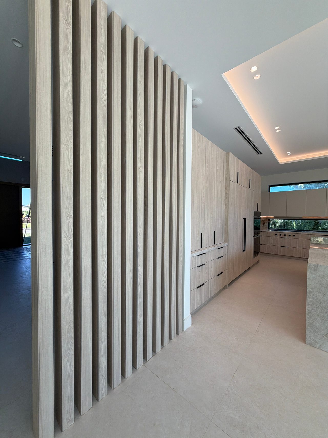 Light wood-grain slat partition with staggered ends beside tall light wood-grain cabinetry, kitchen with island and stone-look tile floor beyond