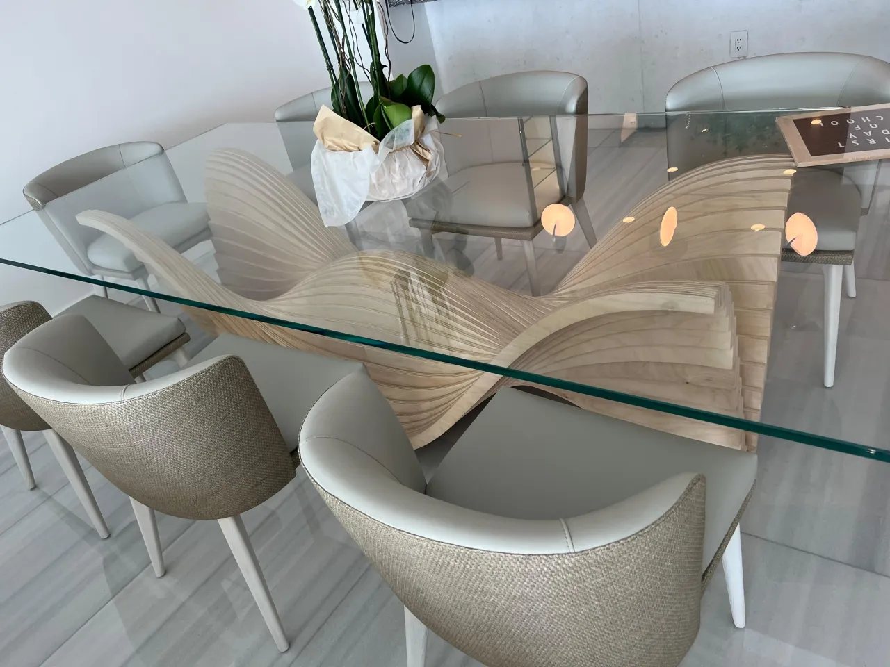 Sculptural wavy wood dining table base carved to resemble undulating waves, topped with a glass tabletop, surrounded by taupe upholstered dining chairs
