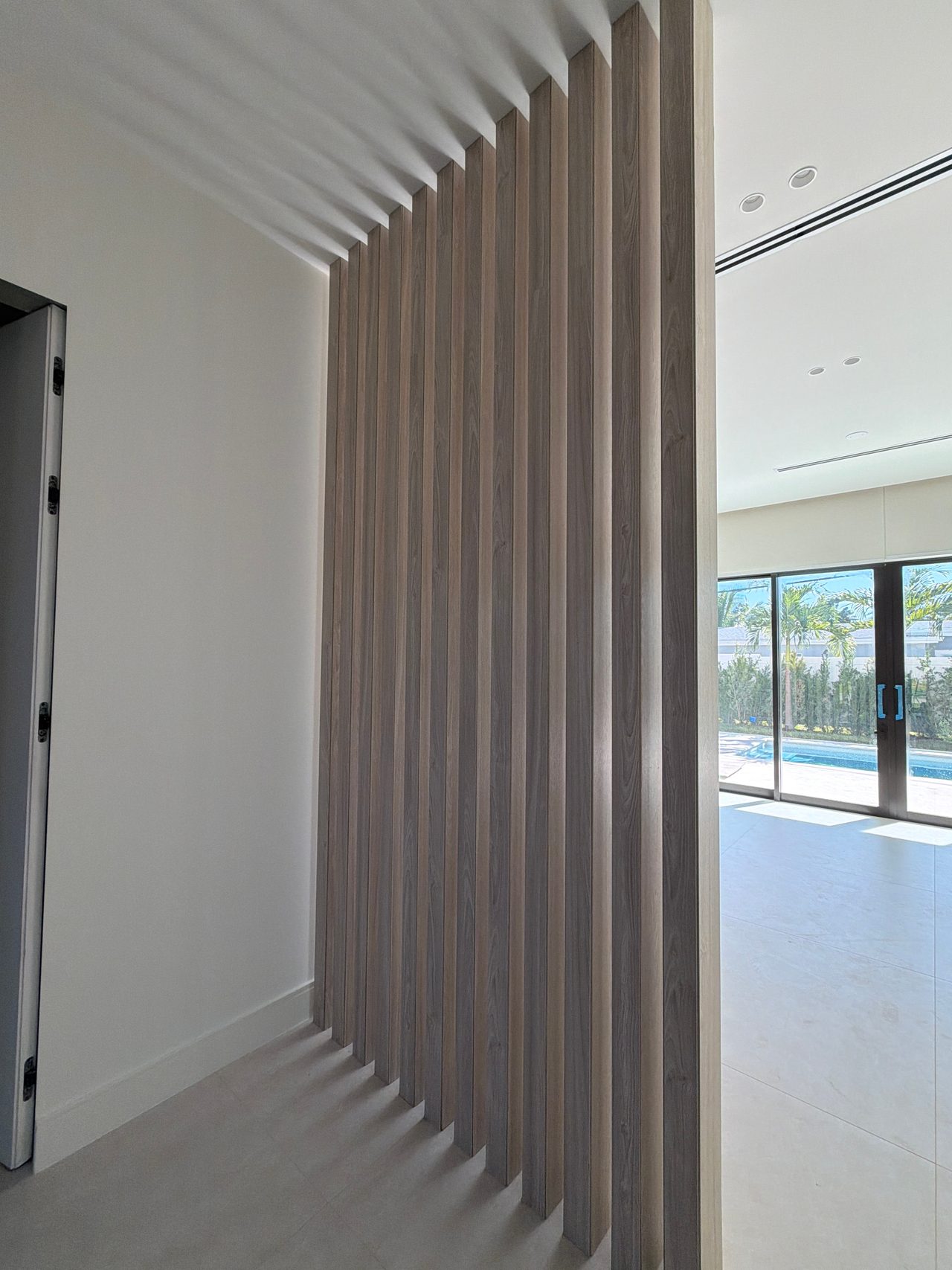 Taupe wood-grain vertical slat screen with staggered ends against a white wall, glass sliders to a pool and palms at right, light tile floor