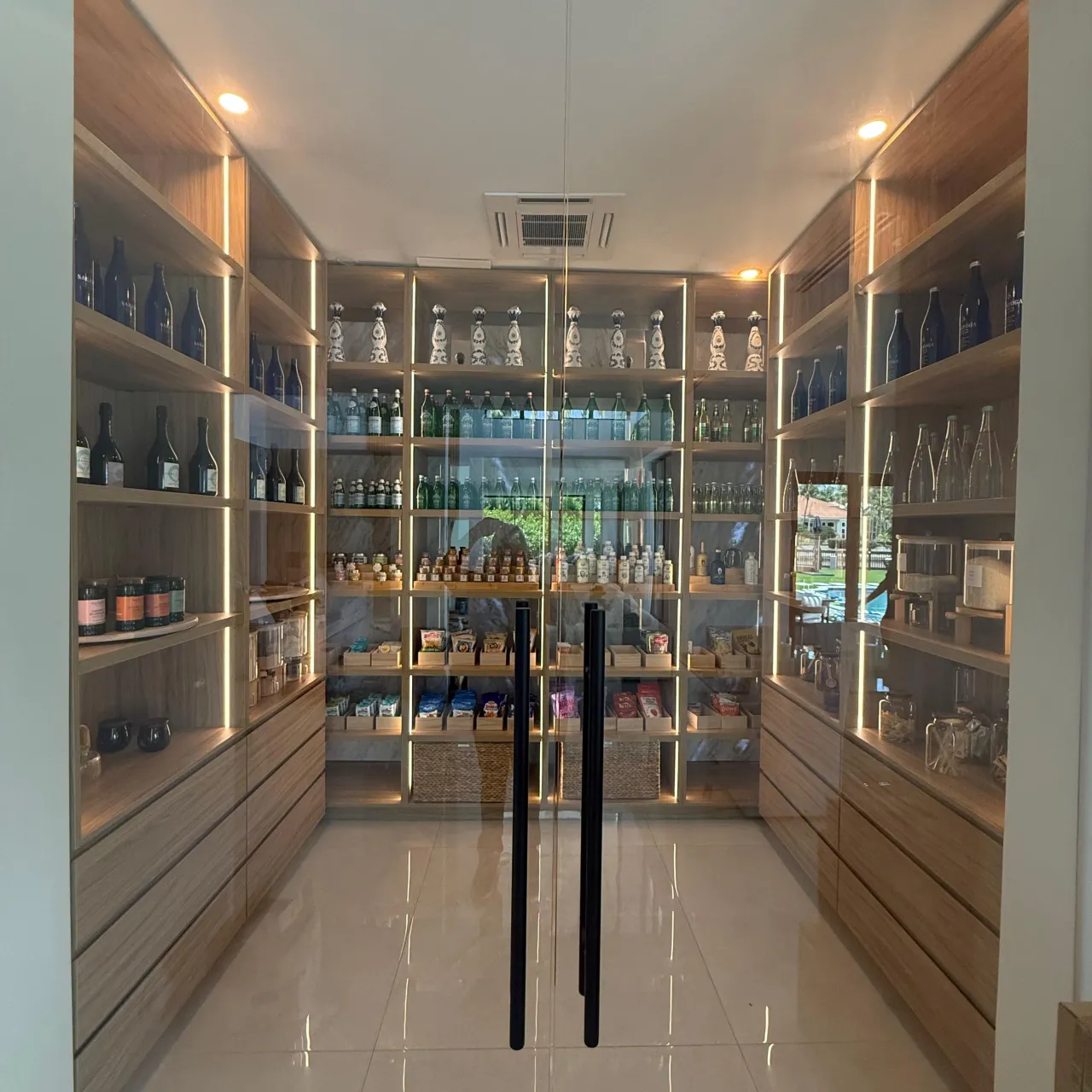 Walk-in pantry lined with wood shelving holding bottles, jars, and packaged goods, with warm LED strip lighting along each shelf edge, glass double doors