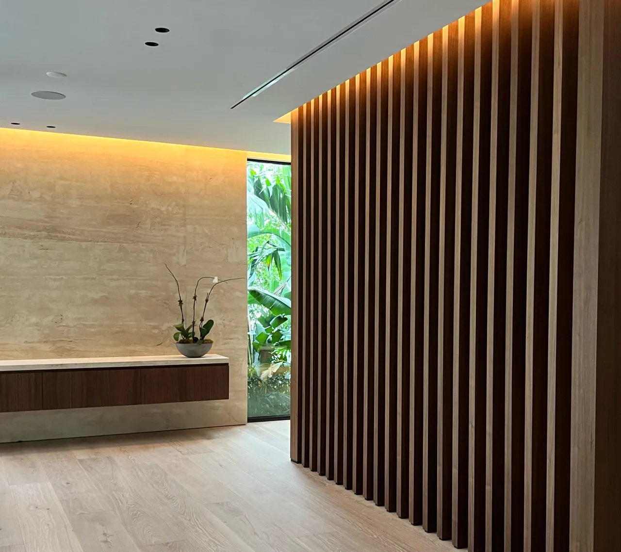 Warm-toned wood vertical slat partition, stone wall at left with a floating wood console and potted plant, glass door to garden foliage, light wood floor