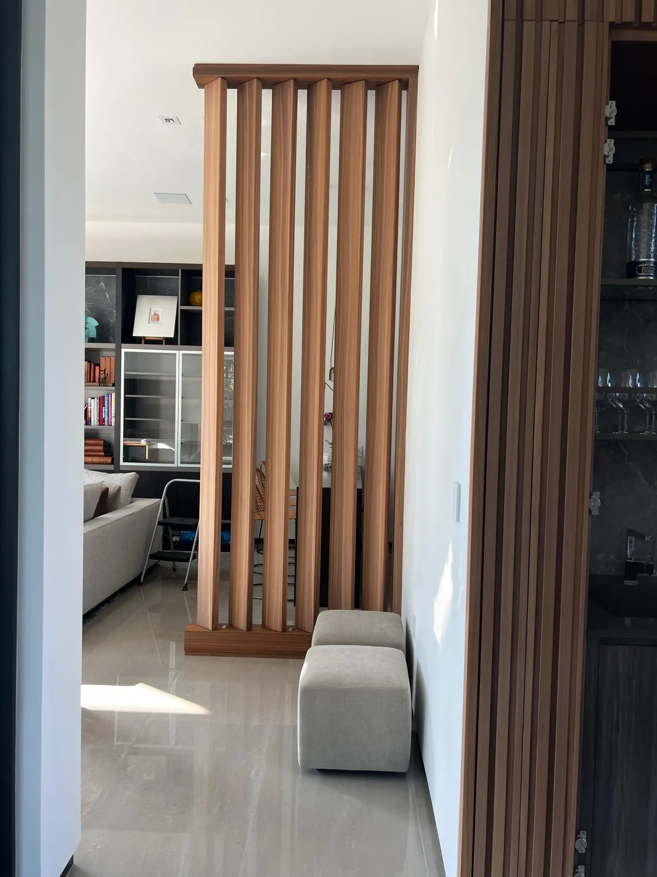 Wood slat divider in a frame between a living room with shelving and a slatted bar cabinet with glassware, gray upholstered pouf on glossy tile floor