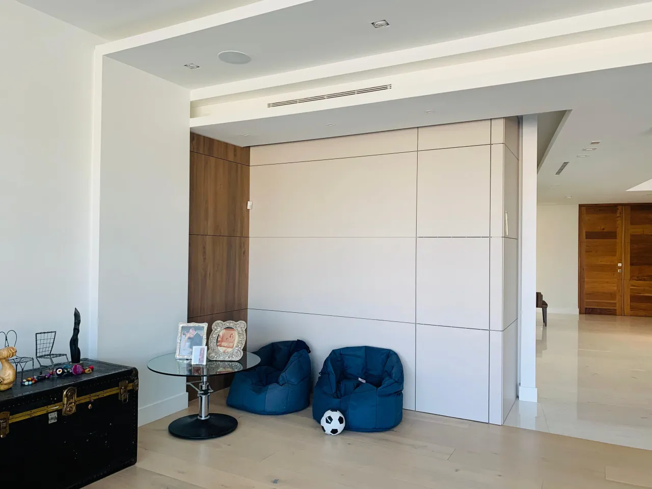 Cream flat-panel wall with recessed reveals and a wood-grain vertical panel at one edge, blue bean bag chairs and a glass side table on wood floor
