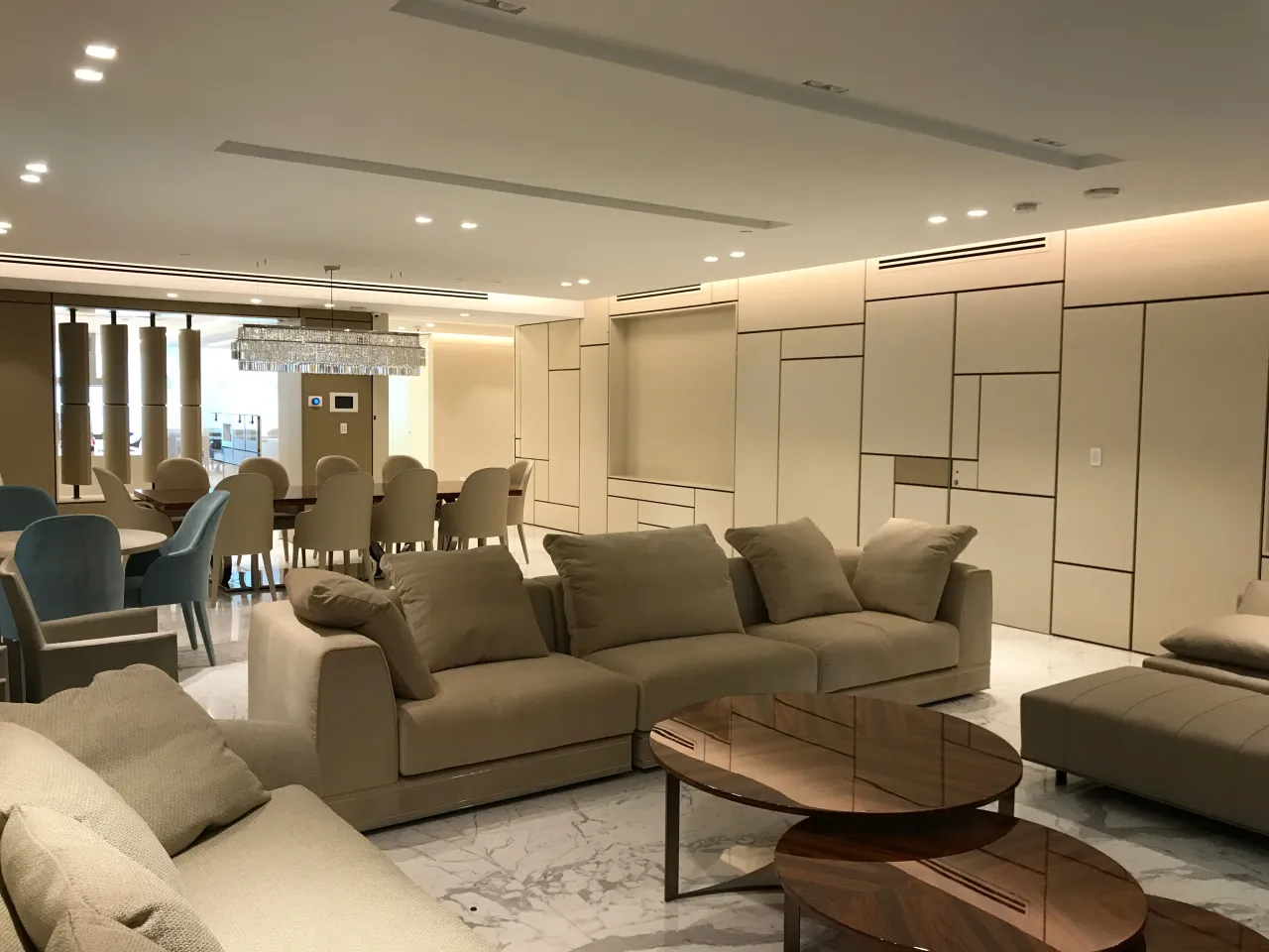 Cream geometric-lined panel wall in an open living and dining area with beige sofas, a dining table with blue chairs, and a crystal chandelier