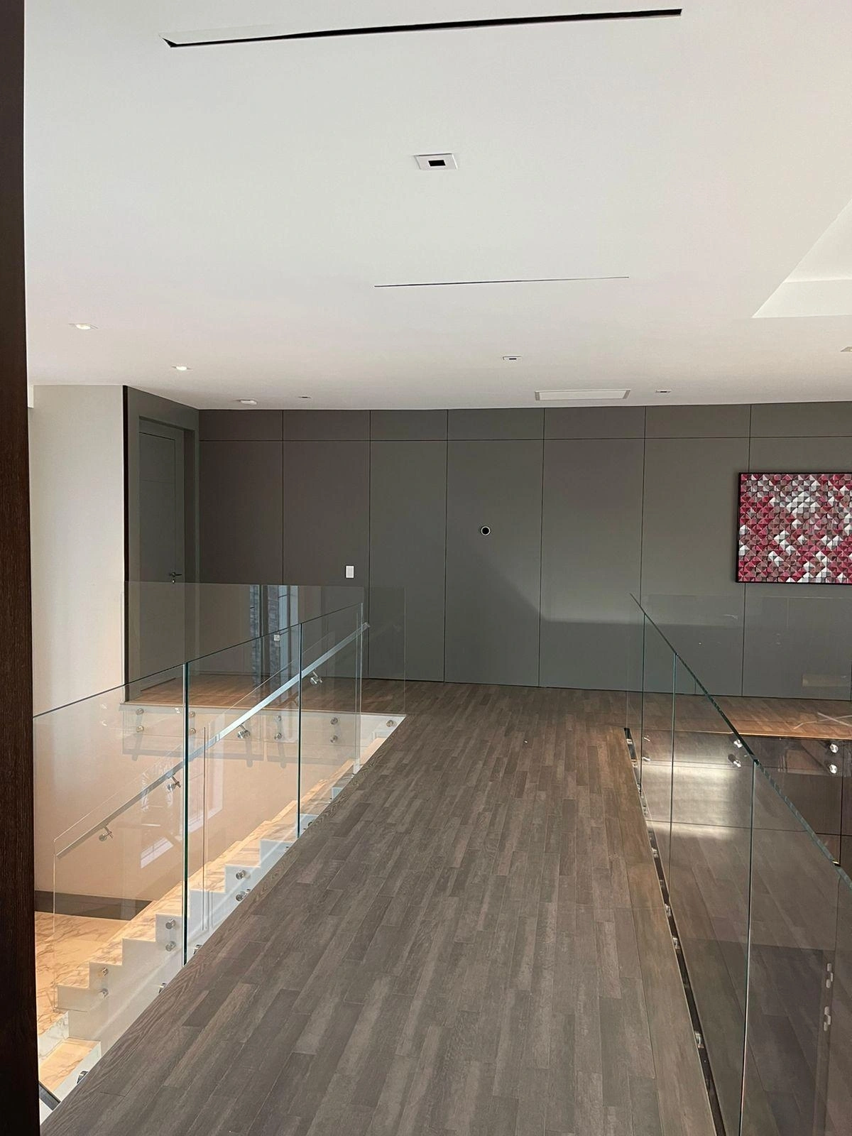 Dark gray flat-panel wall on an upper landing overlooking a stairwell with glass railing, wood-look flooring, small abstract art accent