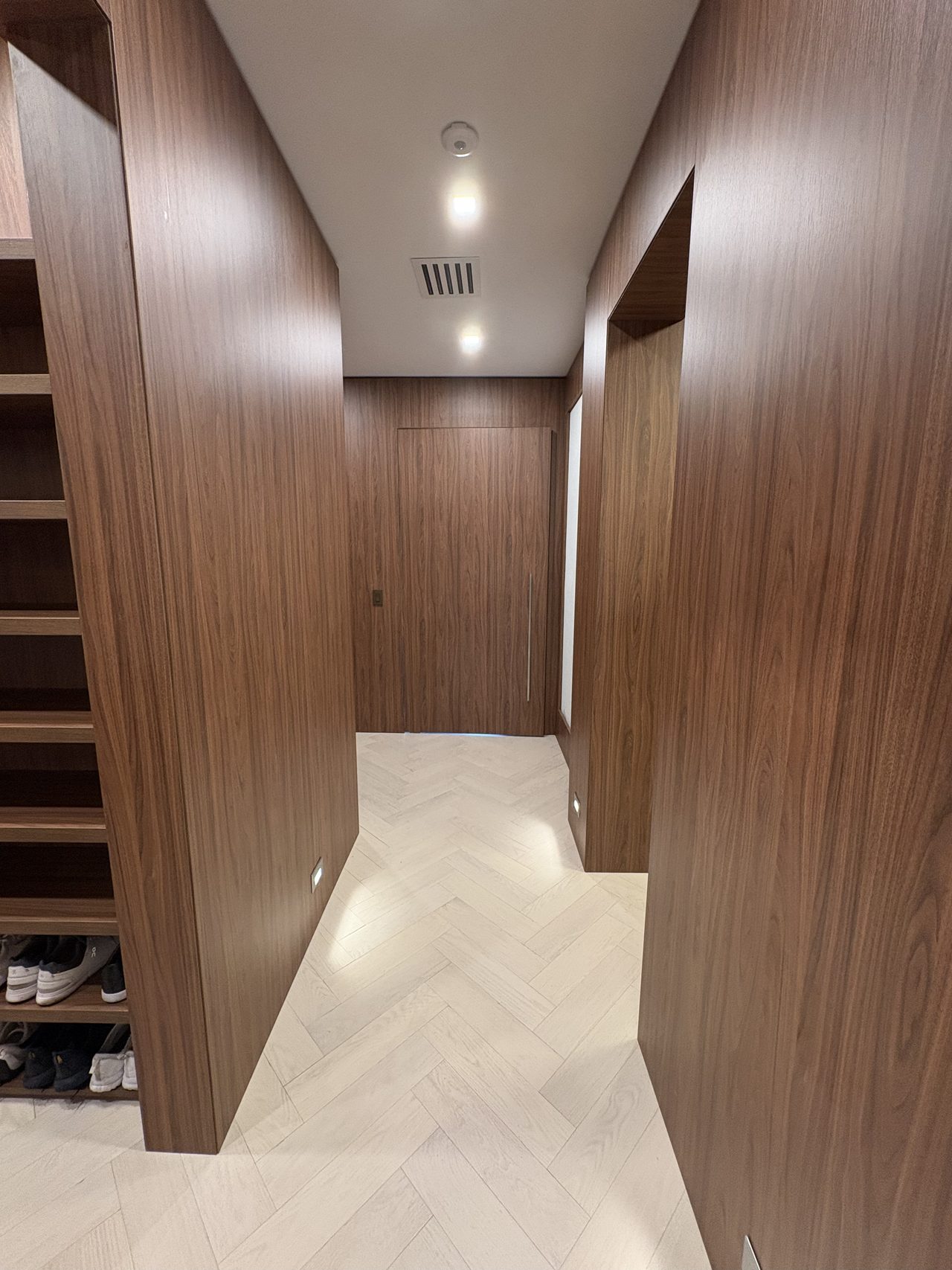 Dark melamine wall cladding corridor leading to a dark wood flush door, with white herringbone-pattern wood flooring and open shoe-shelf cabinet at left