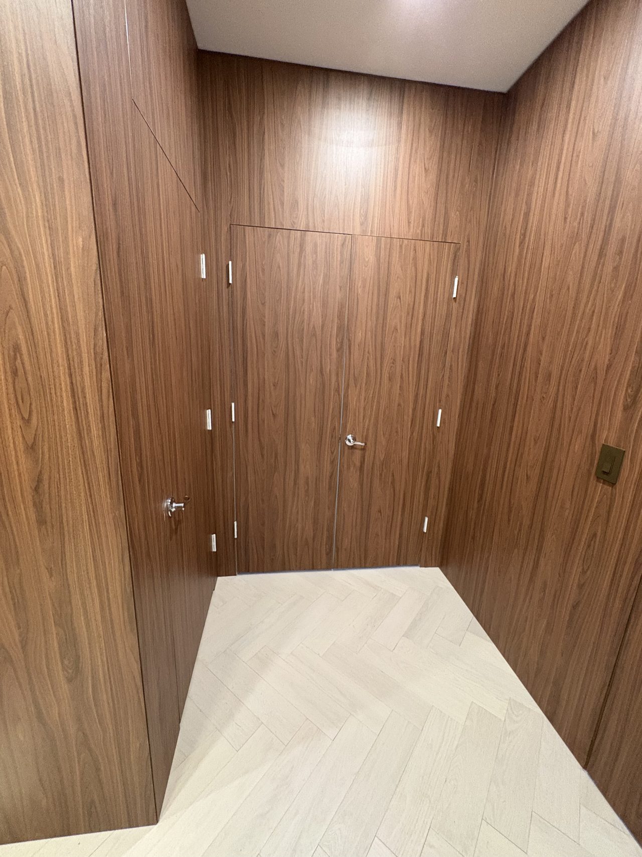 Dark wood-grain melamine-clad corner alcove with a double flush door set, multiple hinge hardware visible, herringbone floor at threshold