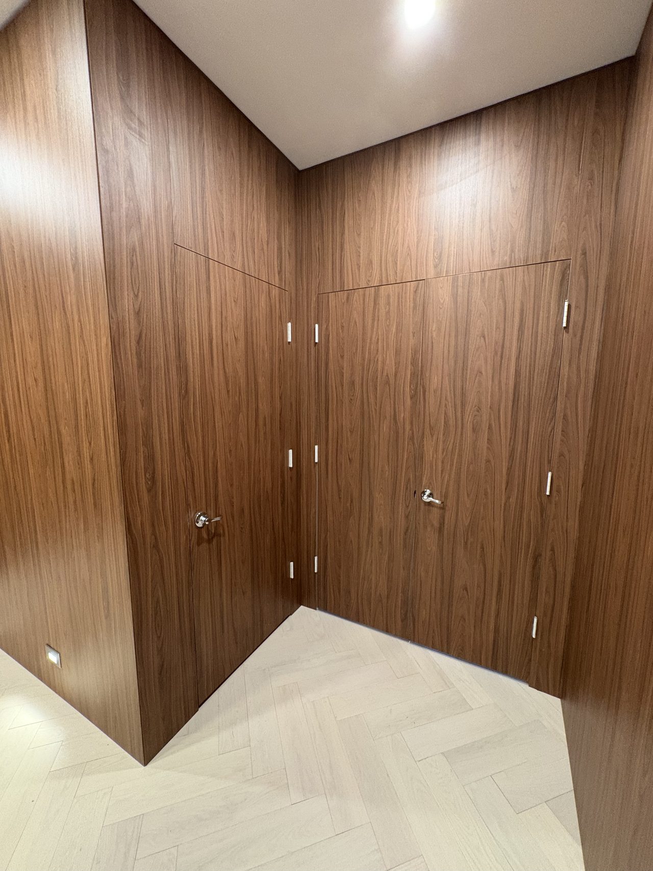 Dark wood-grain melamine-clad corner alcove with a large flush door, chrome door handle, herringbone floor, sloped ceiling detail