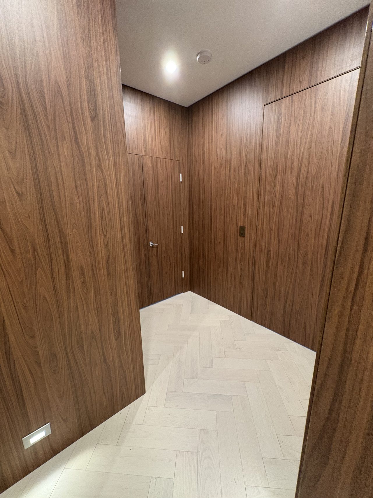 Dark wood-grain melamine-clad corridor corner with two flush doors and a herringbone-pattern floor, recessed ceiling lighting