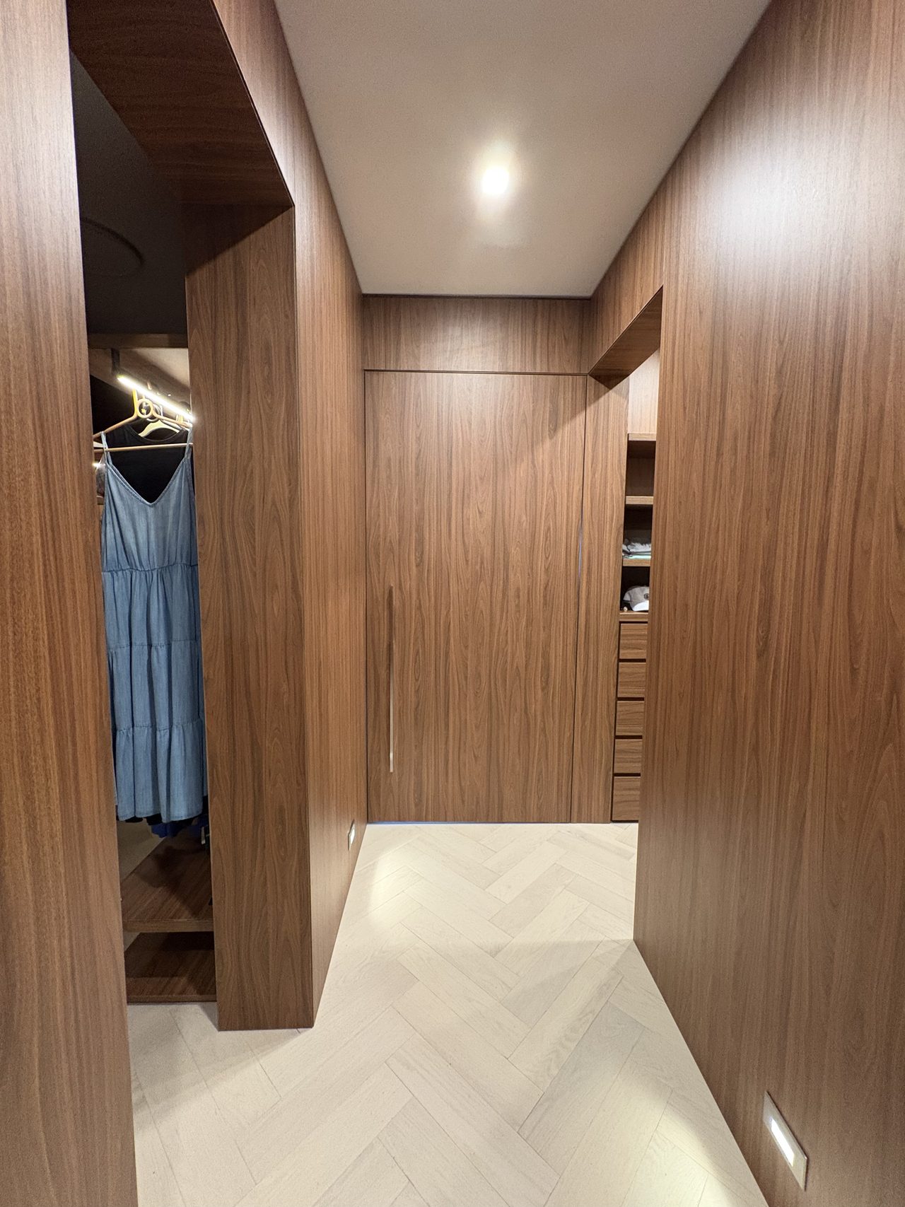 Dark wood-grain melamine wall cladding closet alcove with an open door revealing hanging clothes, wood shelving with drawers, herringbone floor